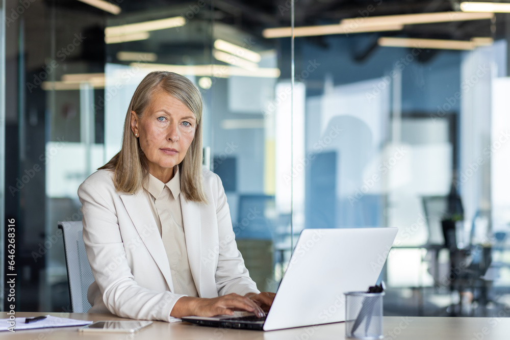 Portrait of serious mature thinking businesswoman, gray haired female boss looking at camera, at workplace inside office, financier with laptop, confident accountant investor. Portrait of serious mature thinking businesswoman, gray haired female boss looking at camera, at workplace inside office, financier with laptop, confident accountant investor.