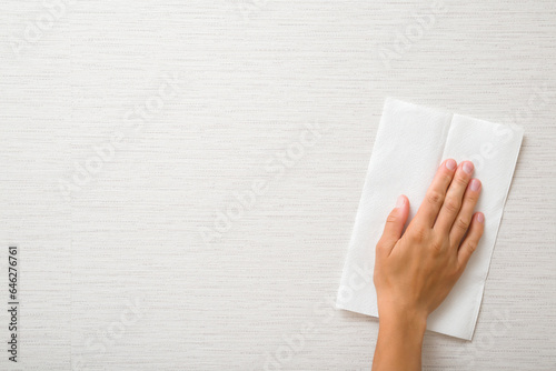 Young adult woman hand holding white dry paper napkin and wiping light gray wallpaper surface. Closeup. Front view. Empty place for text.