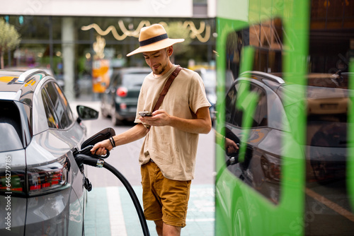 Man in hat plugs a cable in electric vehicle, while standing with phone on a public charging station outdoors. Concept of travel by electric car and green energy for driving