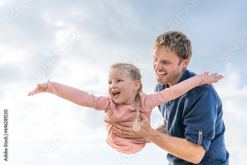 Happy father playing with daughter pretending to be plane under cloudy sky photo