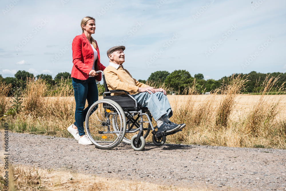 Woman pushing elderly man sitting in wheelchair by field Woman pushing elderly man sitting in wheelchair by field