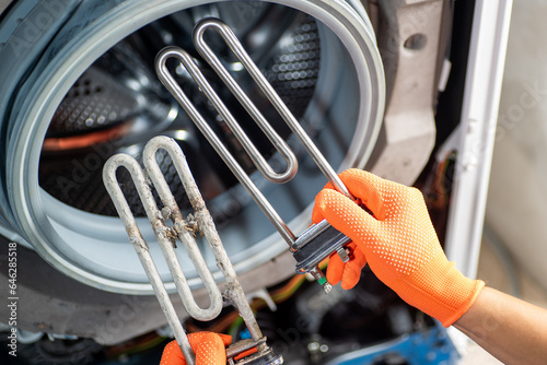 Hands in orange gloves hold two heating elements for a washing machine: one after a long time of use and the other new, against the background of a washing machine. photo