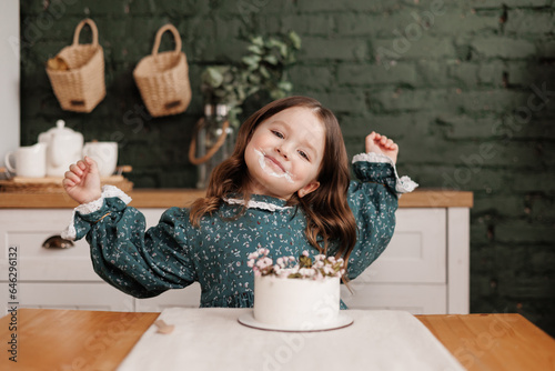 Adorable little child girl wears festive dress takes a bite out of a decorated flowers name cake at a birthday party. Happy smiling kid licks white cream from her dirty face and shows thumb up indoors