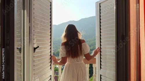 Young brunette woman in a flowing dress opens balcony doors, steps onto the terrace, and revels in a breathtaking mountain sunrise or sunset. Essence of summertime holiday, vacation, nature