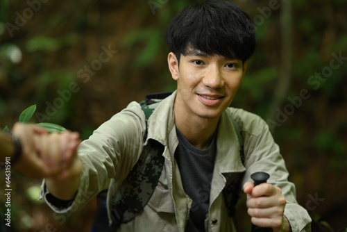 Handsome male tourist helping friend to climb rock in forest. Traveling, trekking and adventure concept © Prathankarnpap