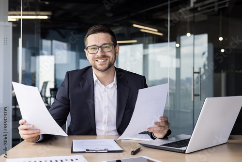 Portrait of a young businessman, an office worker who works in the office on a laptop and holds documents, bills, agreements in his hands. He looks at the camera with a smile