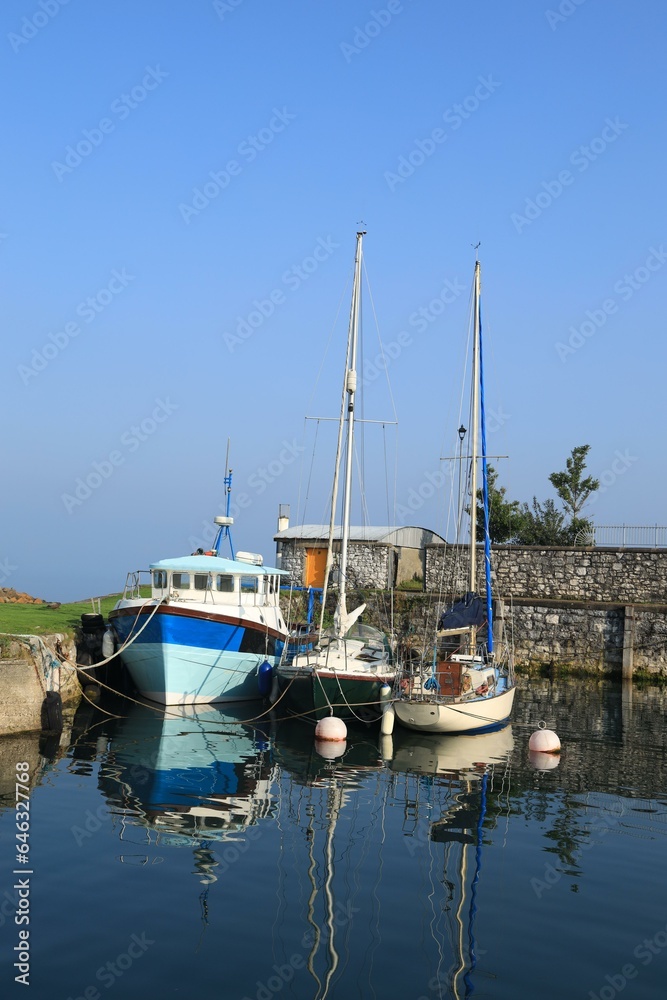Carnlough Harbour in Carnlough, County Antrim, N. Ireland featuring small fishing boats moored in still, reflective tranquil waters against backdrop of blue sky