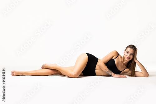 Portrait of young woman sitting on floor, posing in black underwear isolated over white background.