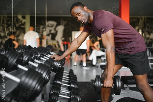 Young African American man sitting and lifting a dumbbell close to the rack at gym. Male weight training person doing a biceps curl in fitness center photo