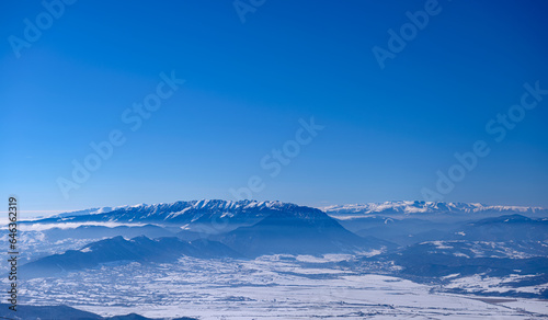 Incredible winter landscape of alpine valley under bright sunny light in frosty morning, beautiful alpine panoramic view of snow capped Piatra Craiului and Fagaras mountains in background, Romania