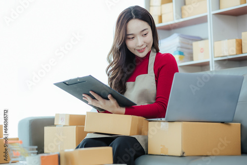 Starting a small business owner happy asian girl Smiling at sales success after checking orders from online shopping store using laptop and smartphone at home office. Concepts of online business and e