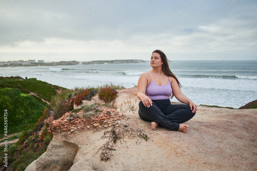 Full figured woman looking away and meditating while sitting at gloomy wild beach
