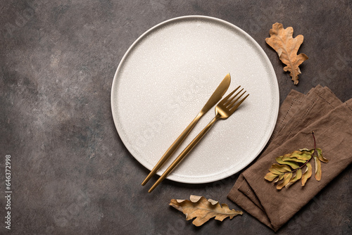 Autumn minimal table setting on brown background. Empty beige plate, golden cutlery, linen napkin and dry autumn leaves. Top view, flat lay.