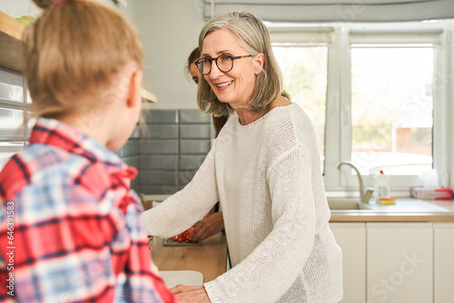 Smiling mature woman in eyeglasses looking at little girl while enjoying time at the kitchen