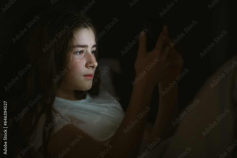 Obraz premium child teenager girl lies in bed in evening or at night and looks into phone. mobile addiction.