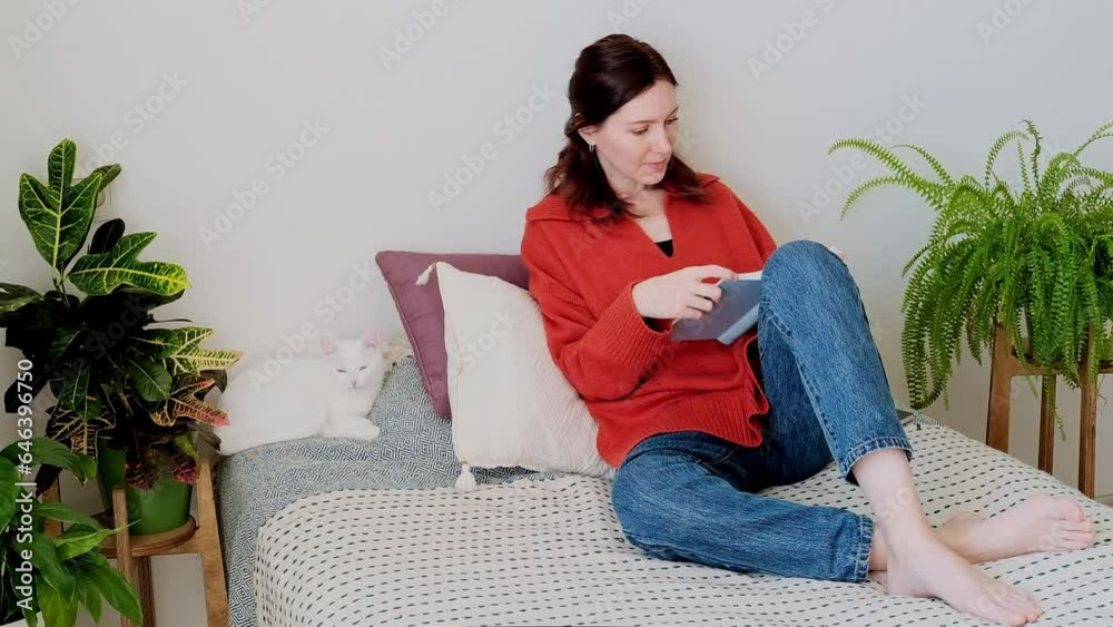 Woman relax sit on sofa with cat reading a book. Attractive woman in a orange sweater sitting on the couch and reading a book at home surrounded by green plants. Woman relax sit on sofa with cat reading a book. Attractive woman in a orange sweater sitting on the couch and reading a book at home surrounded by green plants.