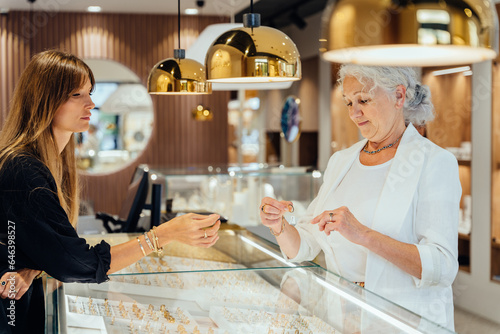 Sale, consumerism, shopping and people concept. Happy grey haired elderly woman buying some jewelry. Blond woman owner, saleswoman of jewelry shop presenting for senior female consumer golden earrings