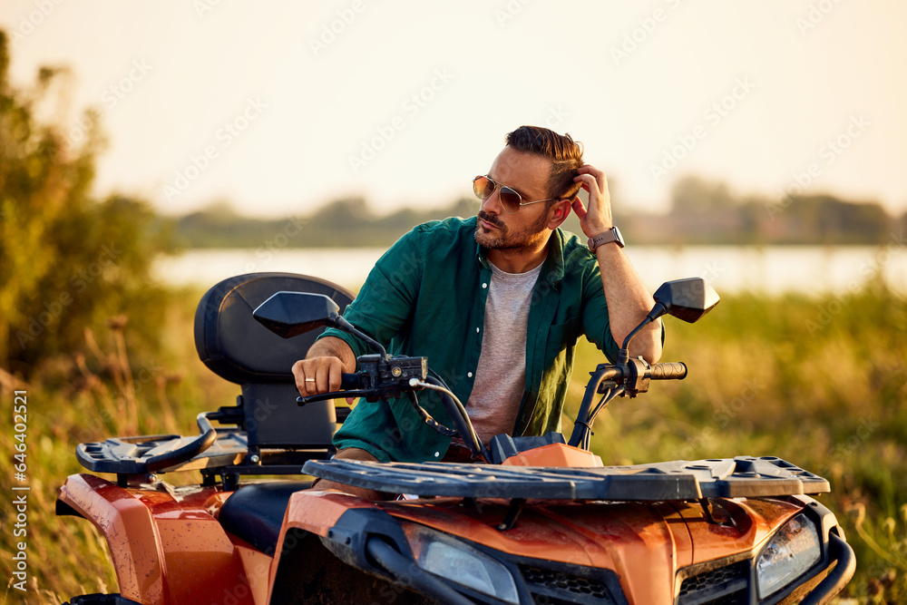An adult man with sunglasses on, sitting on a quad bike and looking around.