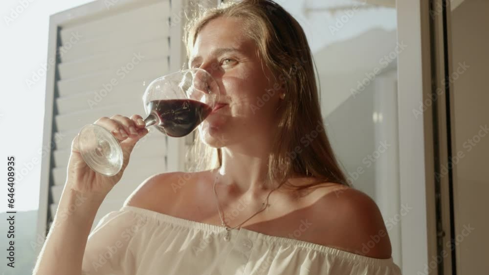 Beautiful young woman stands by an open door on the terrace, delicately holding a glass of red wine, while the sun sets behind mountains. Essence of relaxation and nature's beauty. Beautiful young woman stands by an open door on the terrace, delicately holding a glass of red wine, while the sun sets behind mountains. Essence of relaxation and nature's beauty.