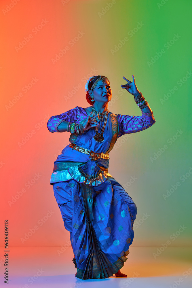 Fototapeta premium Odissi dance. Beautiful mature indian woman in traditional dress dancing indian dance against gradient studio background in neon light. Concept of beauty, fashion, India, traditions, choreography, art