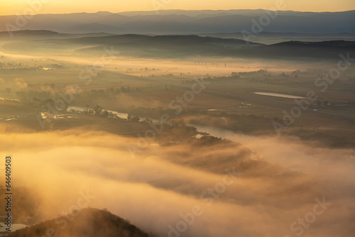 Aerial view of the valley in early morning mist, beautiful in the highlands. Low clouds and fog cover the sleeping meadow. Alpine mountain valley mists landscape at dawn. Serene moment in rural area