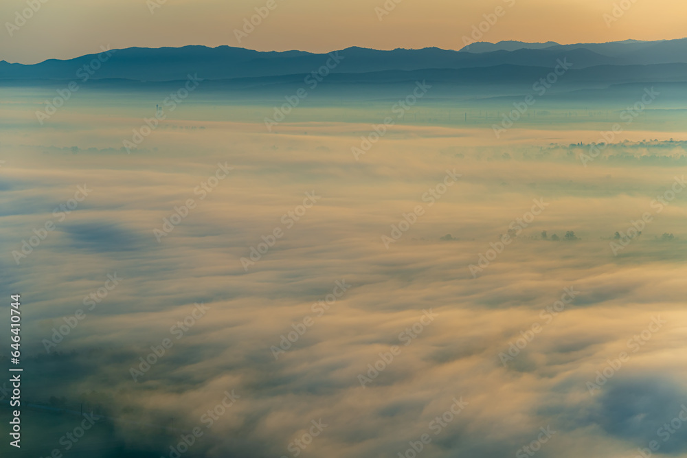 Aerial view of the valley in early morning mist, beautiful in the highlands. Low clouds and fog cover the sleeping meadow. Alpine mountain valley mists landscape at dawn. Serene moment in rural area