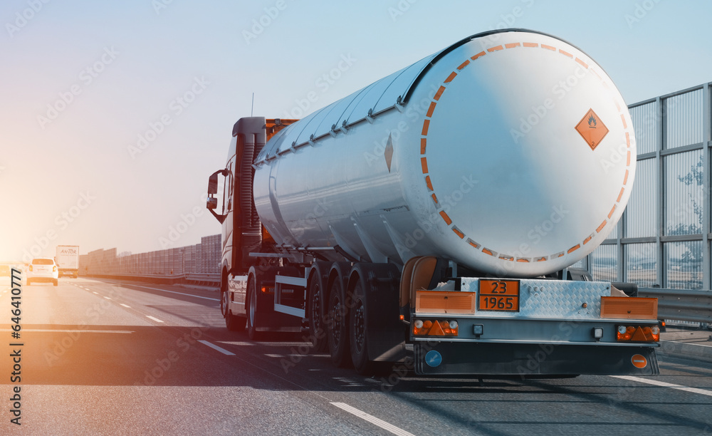 Petrol cargo truck driving on highway hauling oil products. Fuel delivery transportation and logistics concept on a sunny summer evening. Compressed gas carrier truck rear view on a highway.