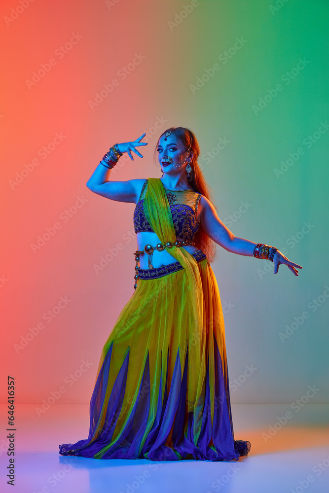 Young beautiful woman in traditional indian dress and makeup dancing against gradient studio background in neon light. Concept of beauty, fashion, India, traditions, lifestyle, choreography, art. Ad