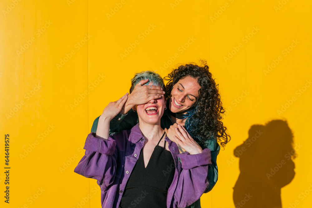 Happy woman covering eyes of lesbian friend in front of yellow wall Happy woman covering eyes of lesbian friend in front of yellow wall