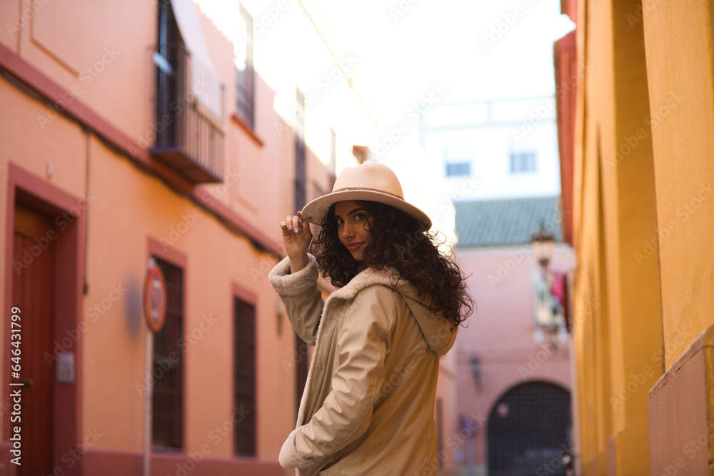 Naklejka premium Young and beautiful Hispanic brunette woman with curly hair wearing a hat and coat for the cold walking in the city of seville while making different expressions and having fun.