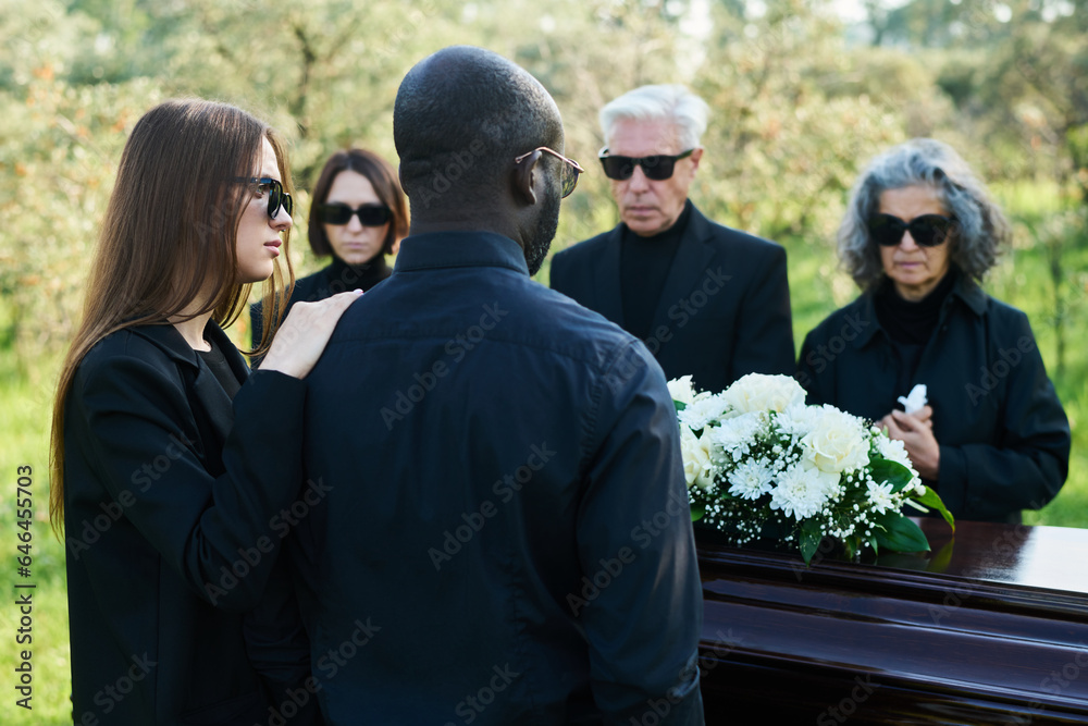 Back view of young woman in mourning attire supporting her husband at funeral while both standing in front of coffin with bunch of white flowers Back view of young woman in mourning attire supporting her husband at funeral while both standing in front of coffin with bunch of white flowers