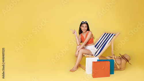 Happy Asian sexy woman dressed in summerwear with shopping bag sitting on beach chair pointing finger isolated on yellow copy space background. Summer vacation sea beach rest concept.