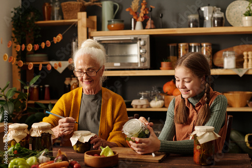 Happy cute girl and her grandmother writing down pickle names on jars while sitting by wooden table after preparation of vegetables for winter