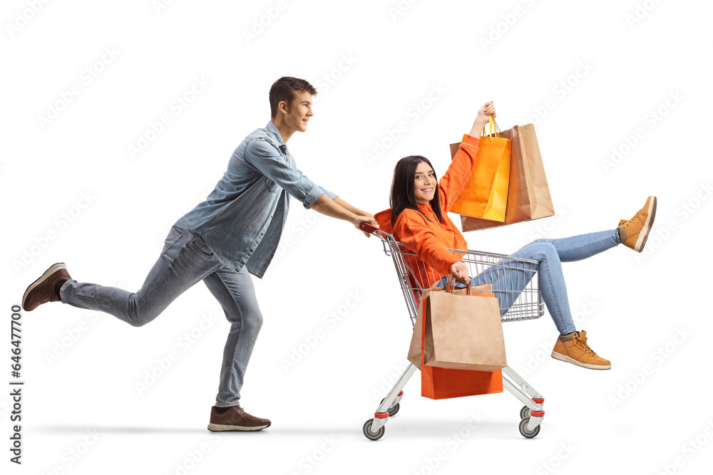 Young man pushing a female with shopping bags inside a shopping cart - obrazy, fototapety, plakaty 
