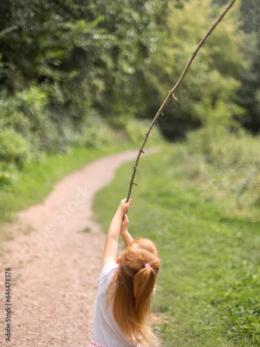 rothaariges Kind spielt mit einem Stock auf einem ruhigen Waldweg – natürliche Außenszene voller Neugier, Freiheit und kindlicher Entdeckerlust.