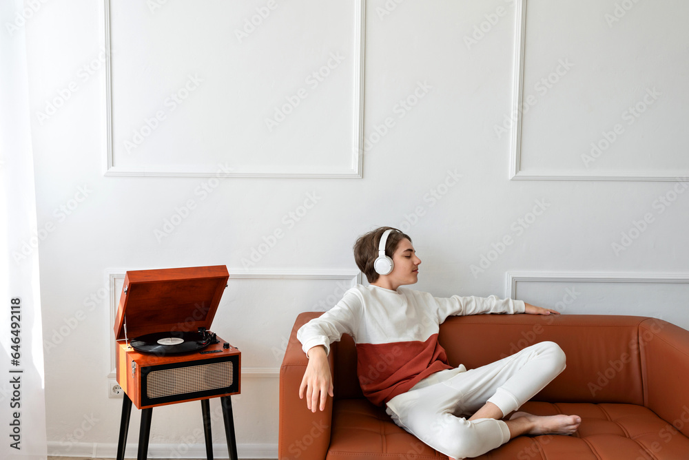 Teenager boy wearing earphones sitting on sofa near wooden retro turntable, chilling and listening vinyl record music. Lifestyle, minimalist home interior, empty white wall background