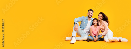 Smiling happy european parents sitting on the floor with child girl, posing on yellow background, panorama, free space