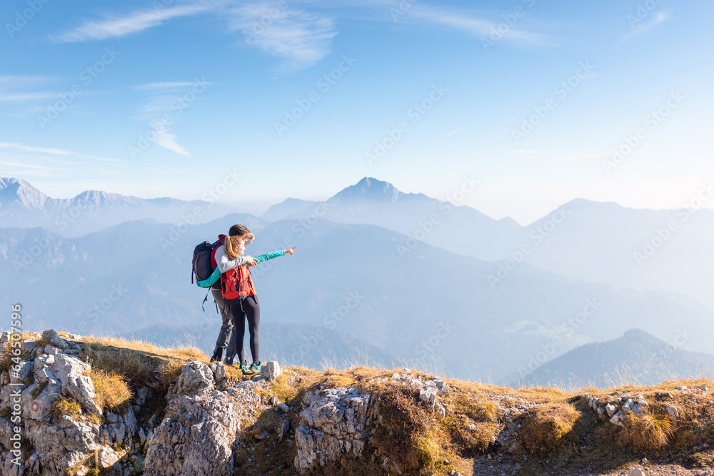 Fototapeta premium Young couple hikers hugging and looking at a fantastic mountain panorama, pointing in the distance on the bright sunny day