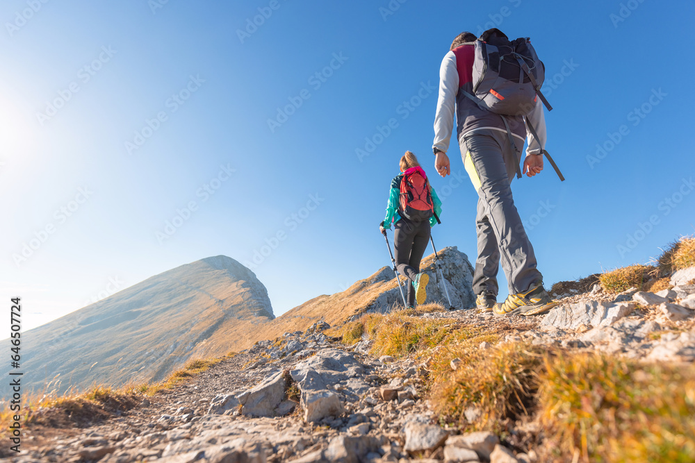 Spectacular view of two hikers walking along the mountain ridge route with the sun on the clear blue sky, aerial shot. Inspiration, nature, and mountaineering concepts. Spectacular view of two hikers walking along the mountain ridge route with the sun on the clear blue sky, aerial shot. Inspiration, nature, and mountaineering concepts.