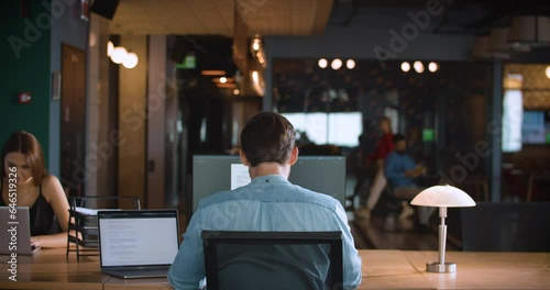 A guy working at his desk in a modern office space, facing a large monitor, with a colleague sitting across from him