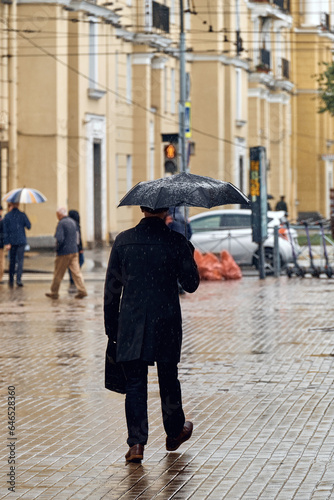 A man in black clothes goes on business on a rainy autumn day. The male hurries to work hiding from the rain under a black umbrella.