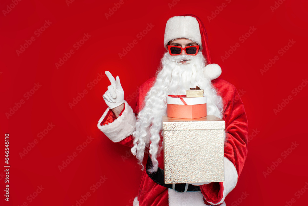 Fototapeta premium santa claus in christmas costume and glasses holds boxes with gifts and shows with his hand to the side