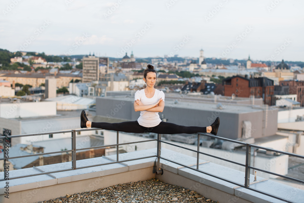 Sportswoman doing side split on roof handrails at sunset