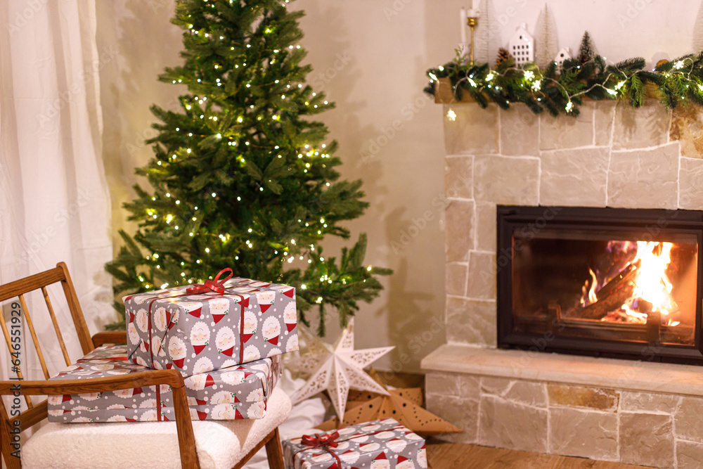 Stylish wrapped christmas gifts on arm chair against christmas tree with festive lights and cozy burning fireplace. Christmas eve, holiday time. Atmospheric living room with xmas presents