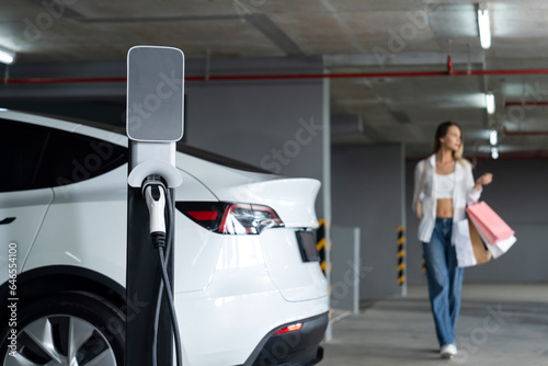 Young woman travel with EV electric car to shopping center parking lot charging in downtown city showing urban sustainability lifestyle by green clean rechargeable energy of electric vehicle innards