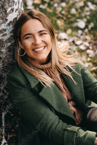 Close up of smiling young woman sitting and leaning on tree while looking at the camera. Portrait of natural beauty woman wearing warm clothes in autumn.