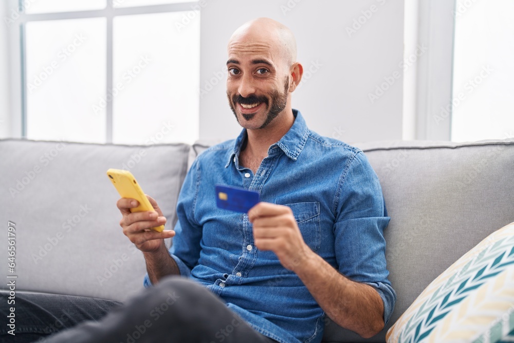 Young hispanic man using smartphone and credit card sitting on sofa at home Young hispanic man using smartphone and credit card sitting on sofa at home