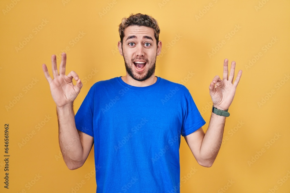 Hispanic man with beard standing over yellow background looking surprised and shocked doing ok approval symbol with fingers. crazy expression Hispanic man with beard standing over yellow background looking surprised and shocked doing ok approval symbol with fingers. crazy expression