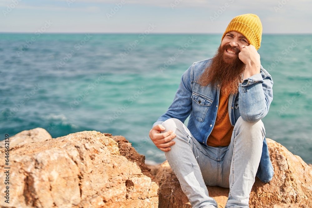 Young redhead man smiling confident sitting on rock at seaside