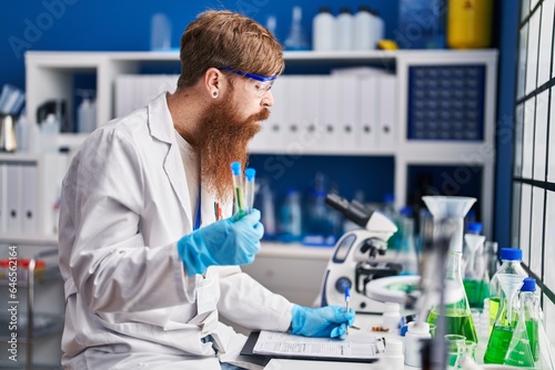 Young redhead man scientist holding test tubes writing report working at laboratory